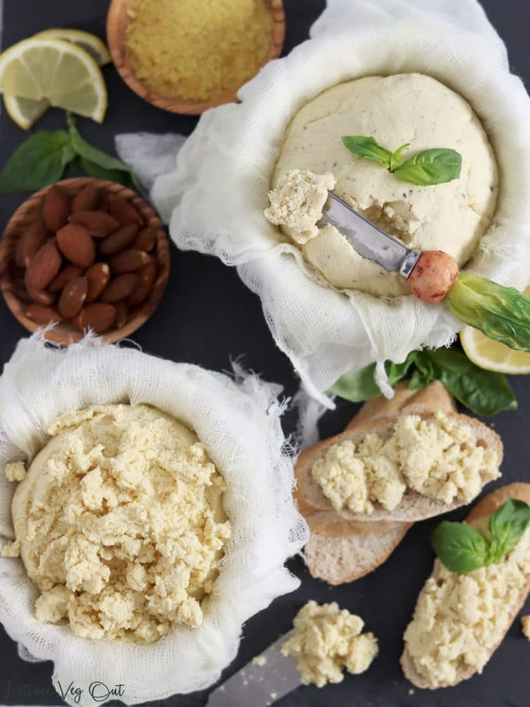Top view of a slate board topped with two balls of almond ricotta cheese, both sitting in cheese cloth. One ball is crumbled and the other mostly whole with a cheese knife in it. Almonds, nutritional yeast, basil, lemon and baguette decorate the board.