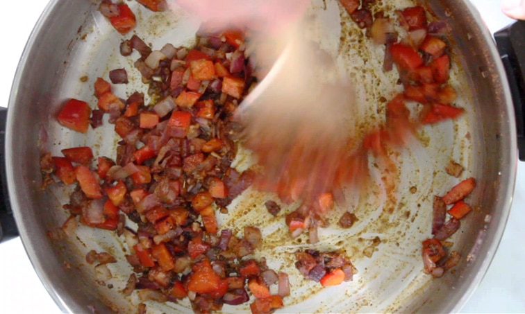 Blurry hand stirring spices onto red bell pepper and red onion in a dry pot.