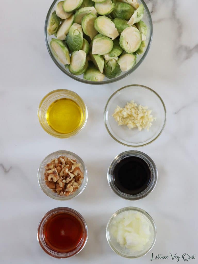 Arrangement of ingredients in glass and wood dishes. From top left moving right then down: Brussels sprouts, olive oil, minced garlic, chopped walnuts, balsamic vinegar, maple syrup, minced onion.