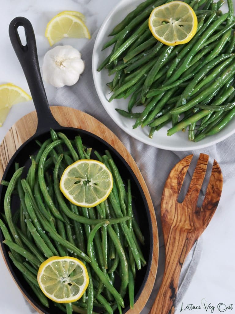 Top view of an oval cast iron pan on an oval wood board, and white plate both filled with bright, roasted garlic green beans topped with lemon slices. Garlic, lemon and a large wood serving fork decorate image.