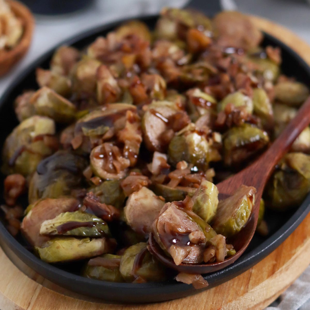 Close up of a small oval cast iron pan sitting on oval wood board and loaded with roasted brussels sprouts with walnuts and balsamic glaze drizzled overtop. Wood spoonful of sprouts on the dish.