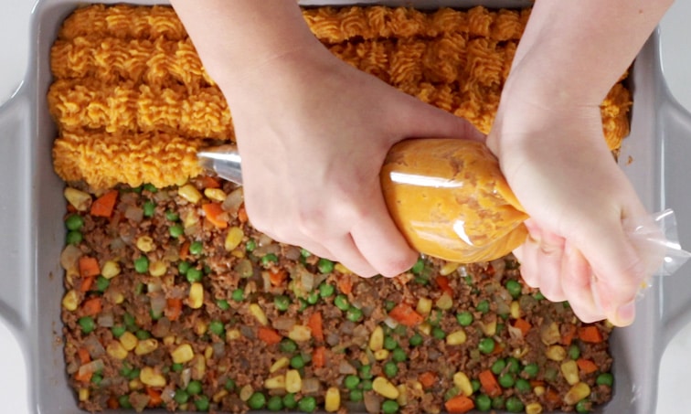 Top view of a baking dish with Shepherd's pie filling in the bottom while hands pipe mashed sweet potato onto the top (piping the 4th row in the center).