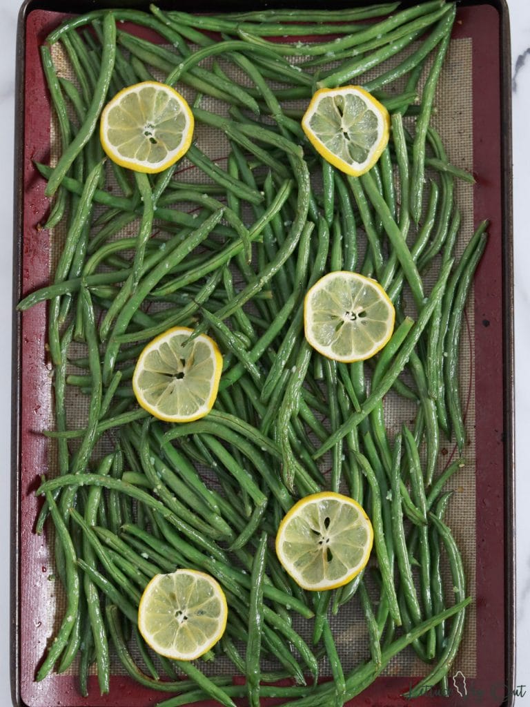 Large baking tray lined with silicone mat loaded with roasted garlic green beans and topped with lemon slices.