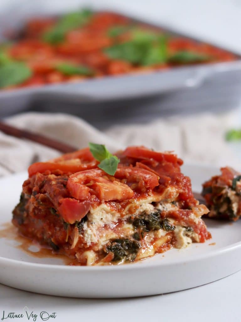 Plate with a large square piece of lasagna, layered with ricotta cheese, spinach and tomato sauce. Grey ceramic dish filled with lasagna sitting on light brown towel in background.