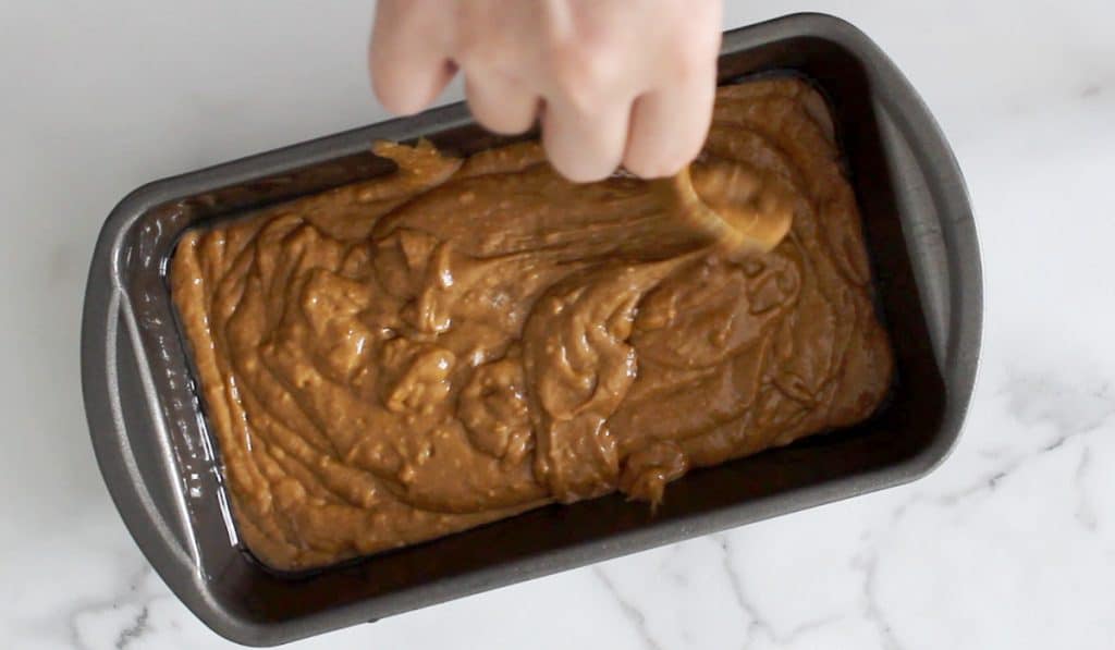 Top view of a loaf pan filled with gingerbread batter while a hand spread batter out with a spoon