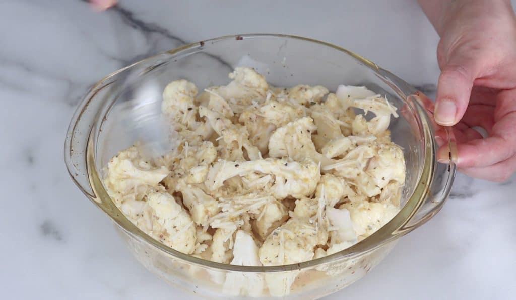 Blurry spoon stirring raw cauliflower florets in a glass dish with a creamy sauce that has shredded cheese and flakes of spices in it.