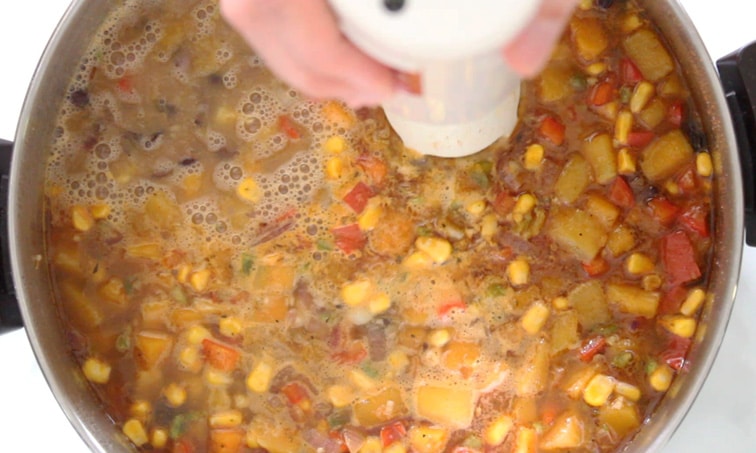 Top view of a pot filled with black bean and squash soup (with corn, onion and bell pepper) which is being pureed by a hand holding an immersion blender into the pot.