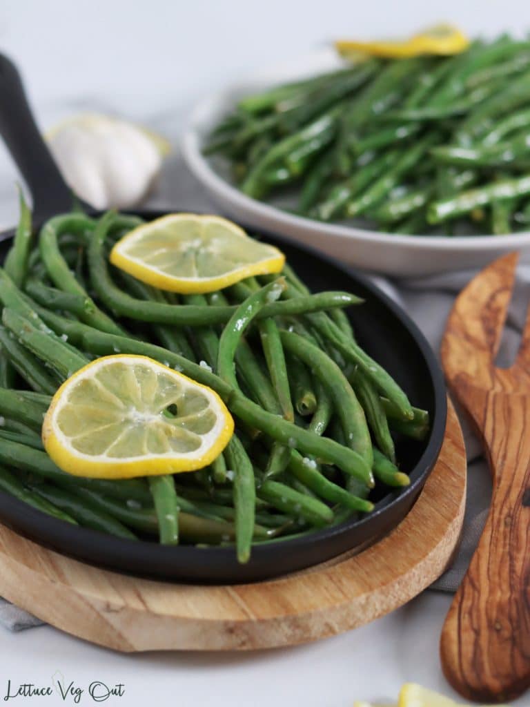 Close up of a small cast iron pan filled with garlic roasted green beans topped with lemon slices. A white plate loaded with green beans and lemon slices in the background.