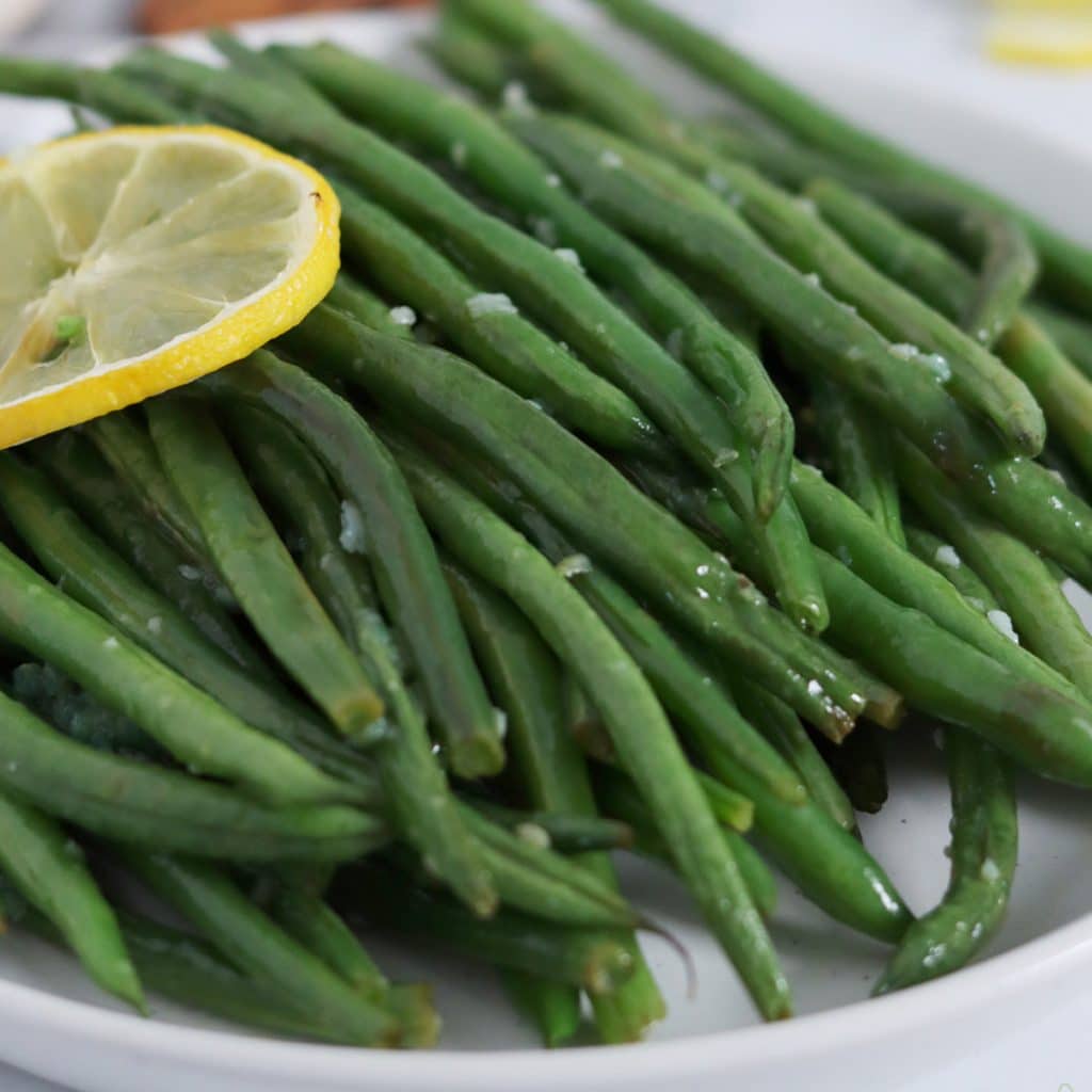 Square cropped close up of a plate topped with a pile of garlic and lemon roasted green beans and garnished with a single slice of lemon.