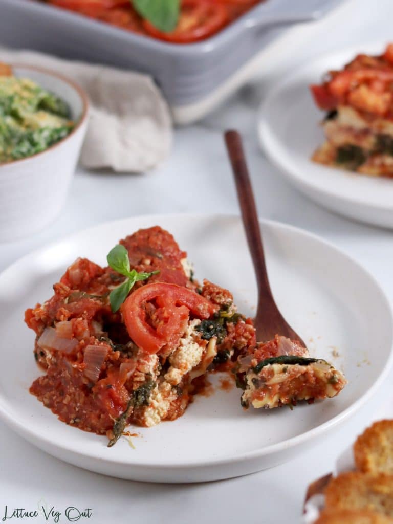 Square of spinach and ricotta lasagna, topped with tomato slices and basil, sitting on a plate with a wooden forkful of lasagna to the right of the slice. Second plate of lasagna, bowl of Caesar salad and ceramic lasagna dish sit cropped out and blurred in background.