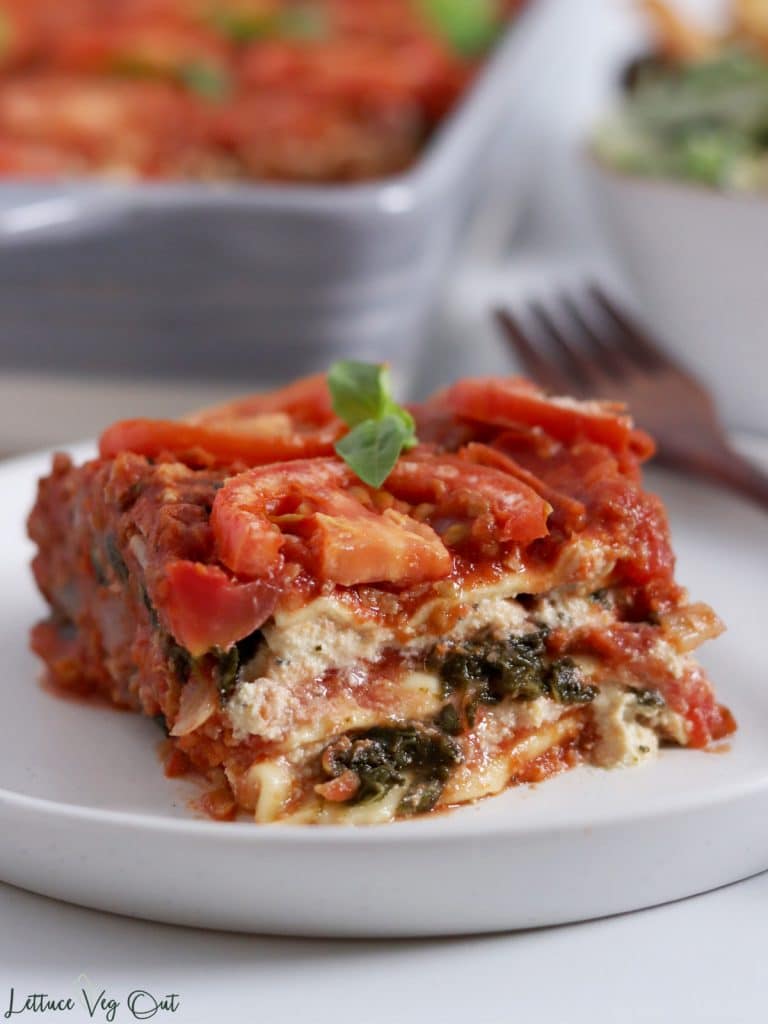 Close up of a piece of vegan spinach and ricotta lasagna on a plate, topped with tomato slices and basil. Wood fork on the plate with a blurry lasagna dish and bowl of Caesar salad in the background.