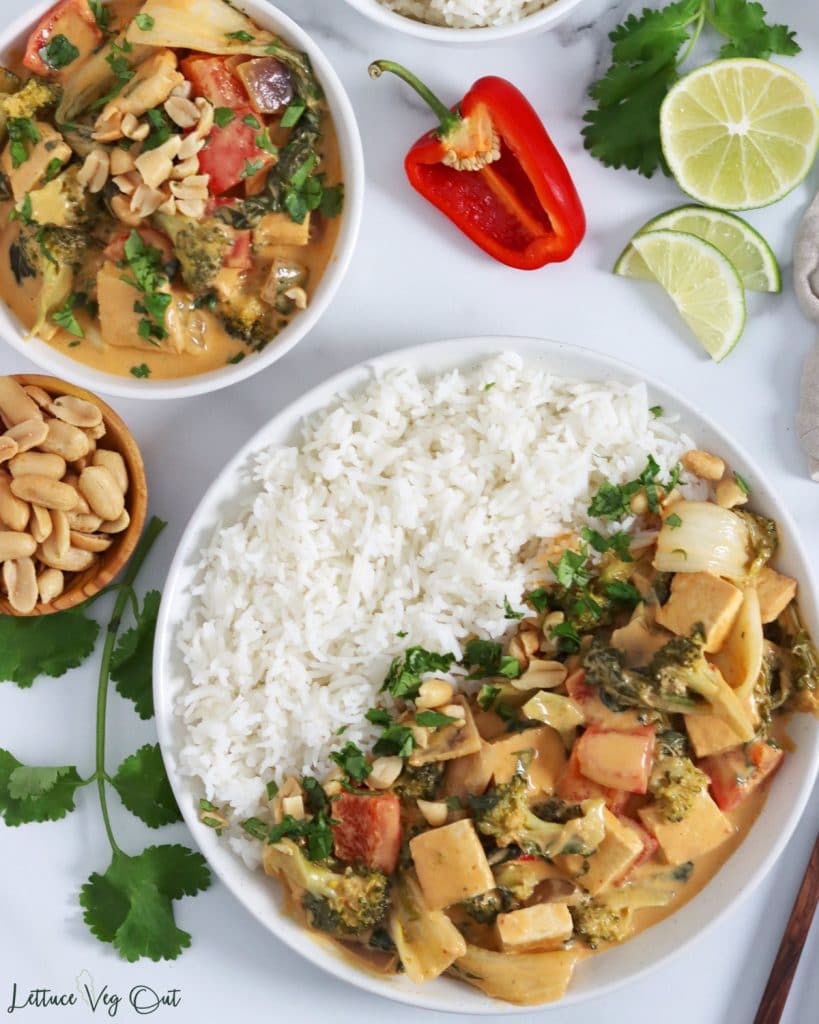Top view of a plate half filled with rice and the other half with peanut butter red curry (with tofu, bok choy, bell pepper and broccoli). Small bowl of curry in top right corner and image is decorated with cilantro, lime wedges, mini red peppers and a small wood dish of peanuts.
