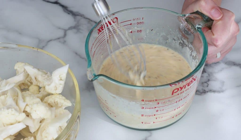 Blurry whisk that is whisking a creamy sauce in a glass measuring cup with a hand griping the handle of the cup.