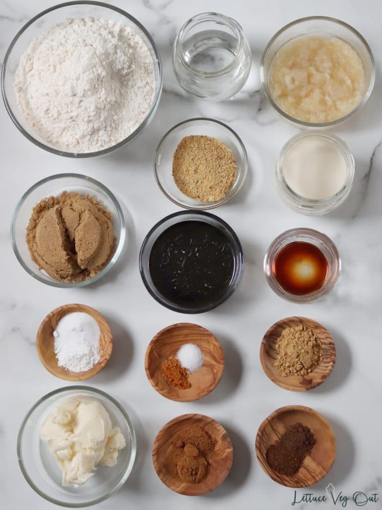 Arrangement of ingredients in glass and wood dishes. From top left working right then down: flour; water; applesauce; ground flaxseed; milk; brown sugar; molasses; vanilla extract; baking powder and soda; salt and ground mace; ground ginger; butter; ground cinnamon and nutmeg; ground allspice and cloves.