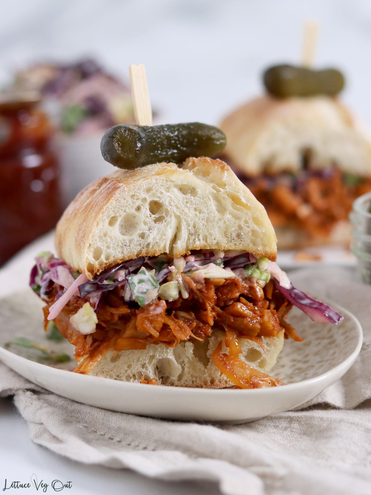 Close up of a small pulled jackfruit sandwich with creamy coleslaw on it, topped with a pickle. A second sandwich and jar of BBQ sauce sit behind the first sandwich.