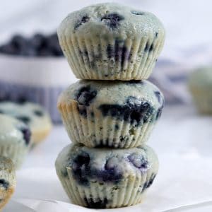 Close up of a stack of 3 blueberry muffins on a piece of parchment paper with other muffins and a bowl of blueberries in the background.