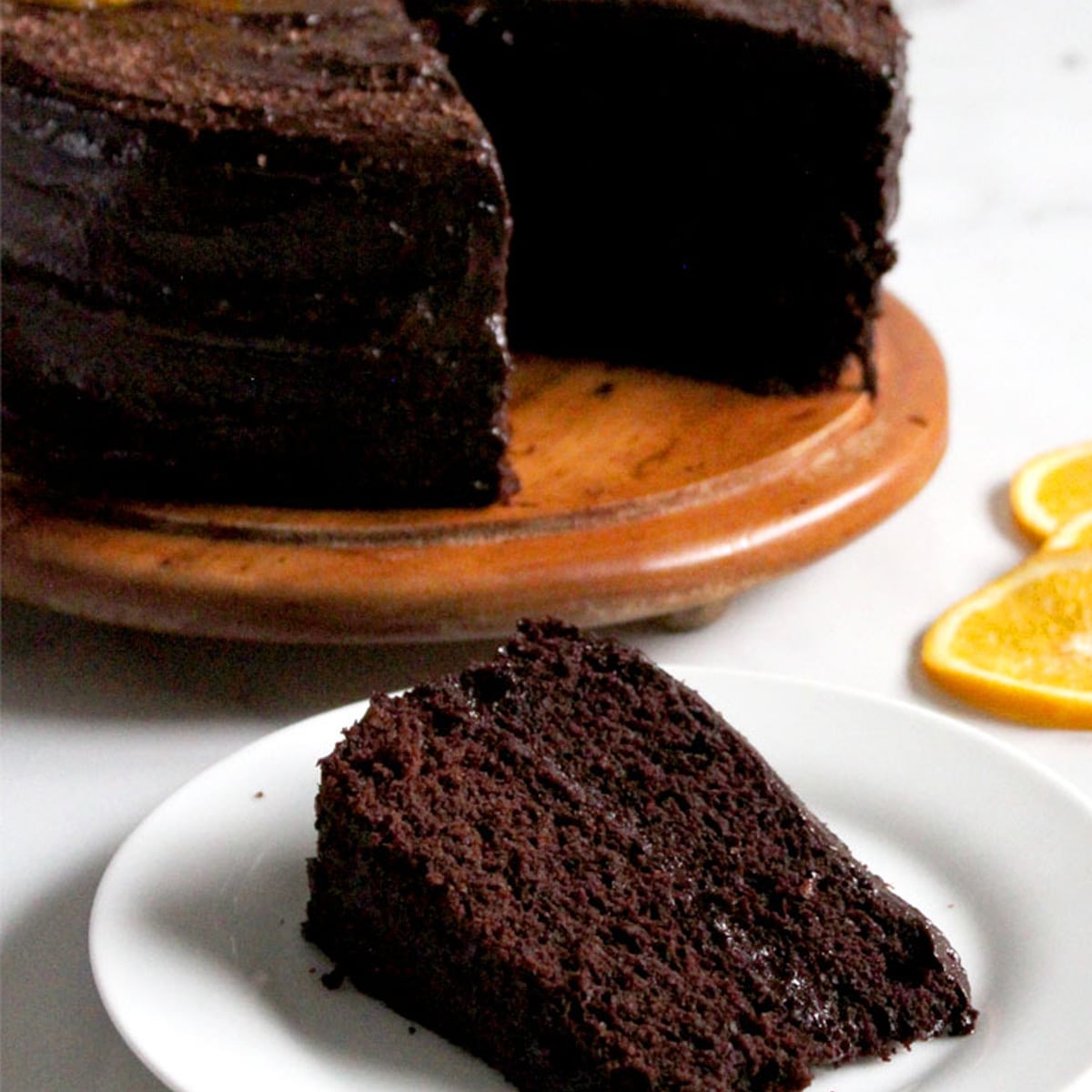 Close up of a slice of chocolate orange cake on a plate in front of a wood cake board with the double-layer cake on it.
