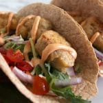 Close up of a taco filled with breaded tempeh, tomato, onion, lettuce and orange Baja sauce.