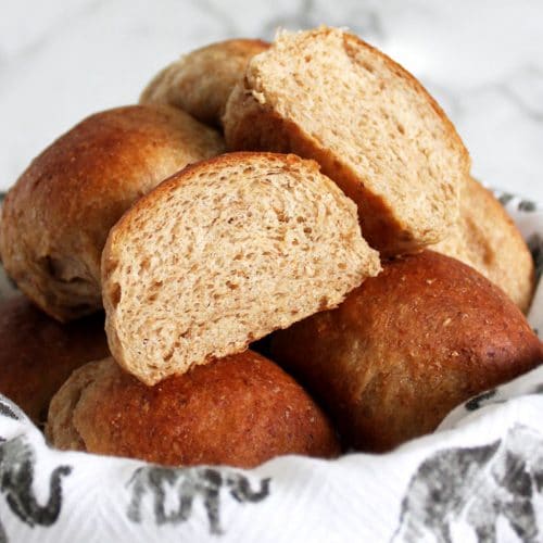 Pile of whole wheat rolls in a bowl with one roll cut open to show the inside.