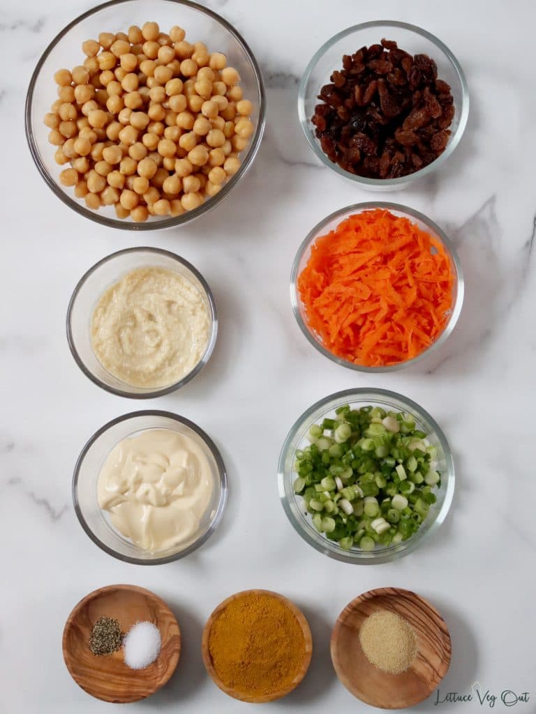 Arrangement of ingredients. From top left moving right then down: chickpeas, raisins, hummus, grated carrot, mayo, green onion, salt and pepper, curry powder, garlic powder.