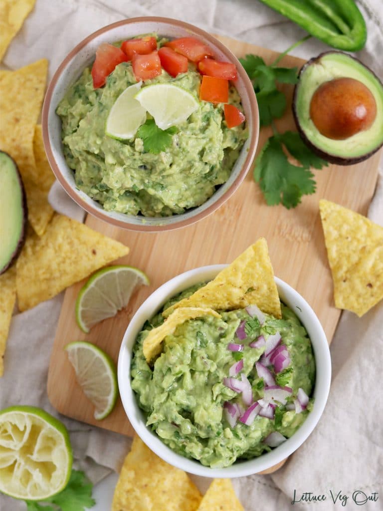 Two bowls of guacamole on a wood board with corn chips, lime and tomato garnish.
