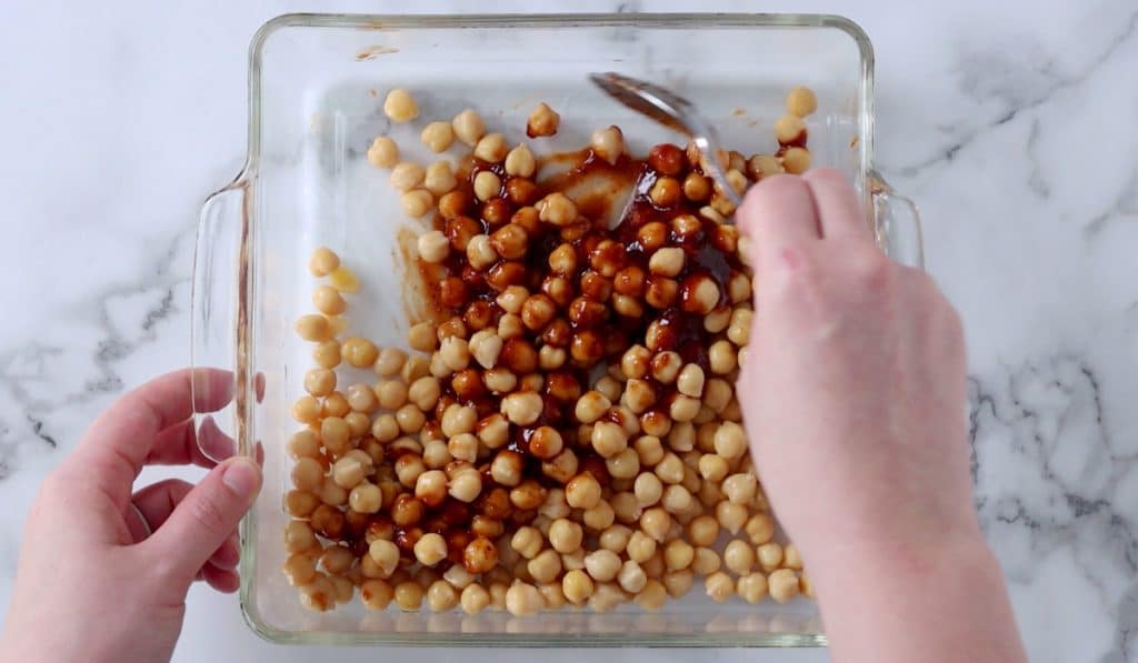 Stirring barbecue sauce and chickpeas in a square glass baking dish.
