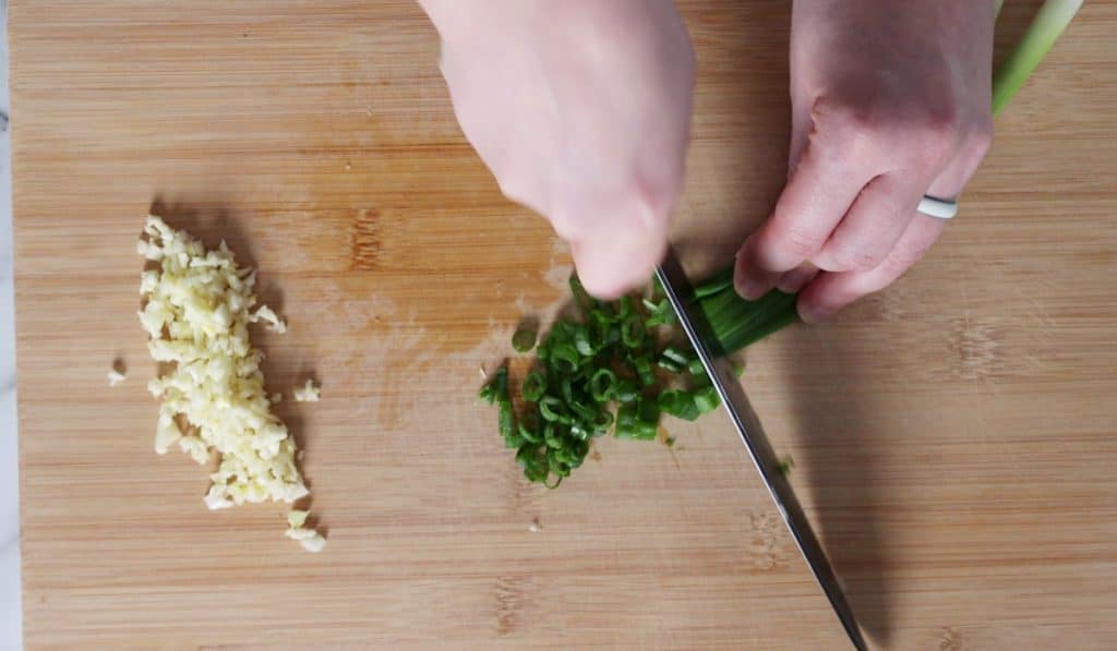 Hands chopping green onion next to minced garlic on wood board.