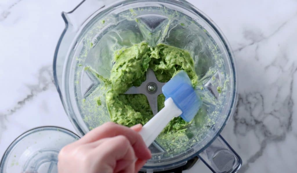 Hand scraping partially-blended avocado sauce from edges of blender.