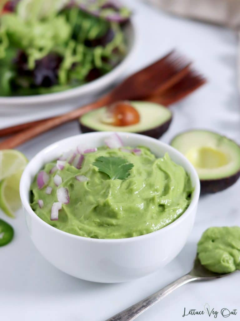 Bowl of avocado sauce with a halved avocado and plate of salad in the background. Text at top reads "Vegan Avocado Sauce".