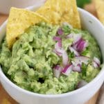 Close up of a bowl of guacamole garnished with red onion and two corn chips.