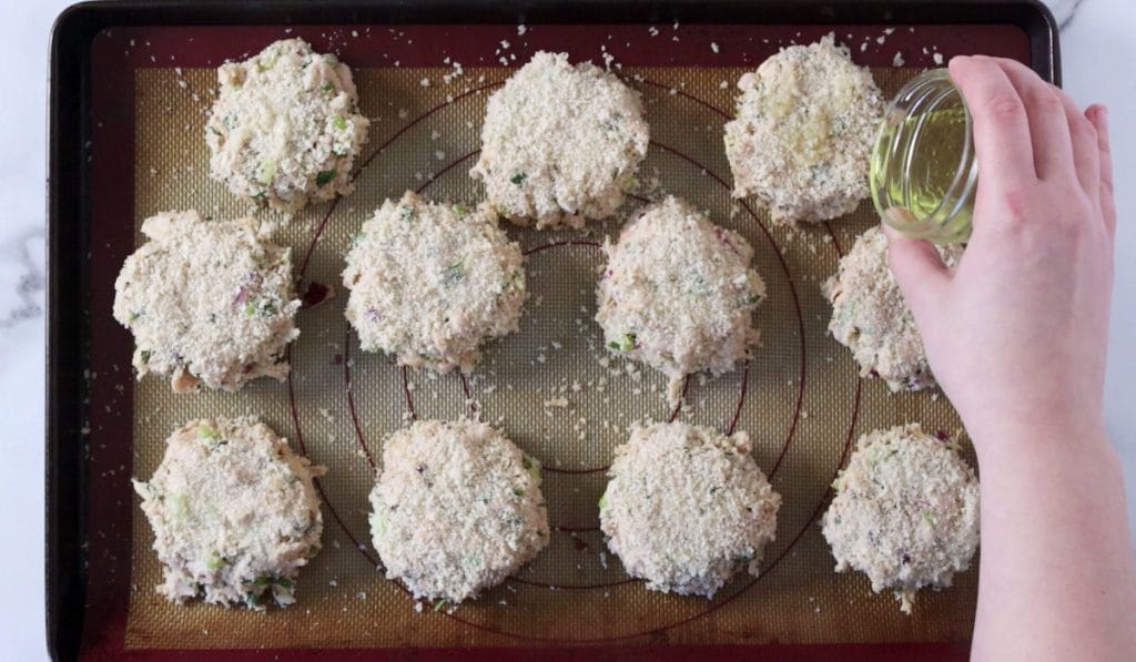 Baking tray with breaded crab cakes on them. Hand drizzling oil onto crab cakes.