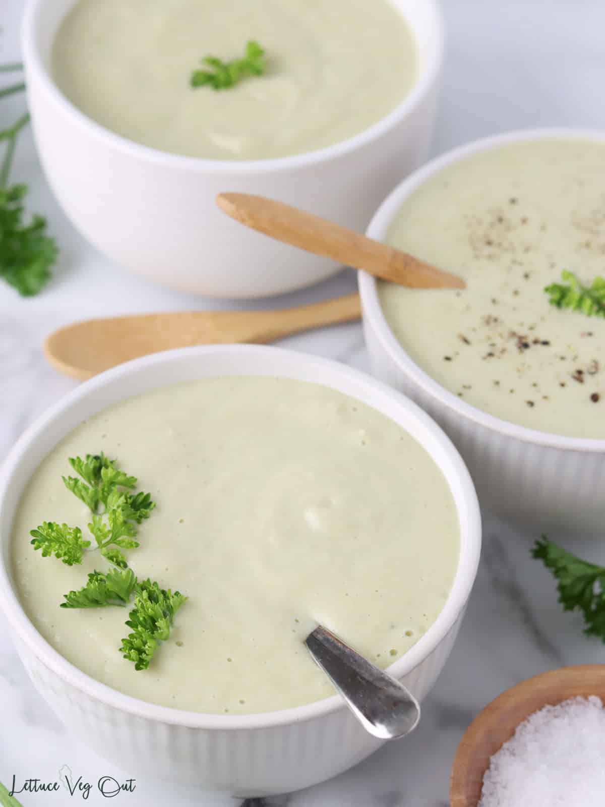 White bowl of creamy celery soup with parsley garnish and two other bowls blurred in background.