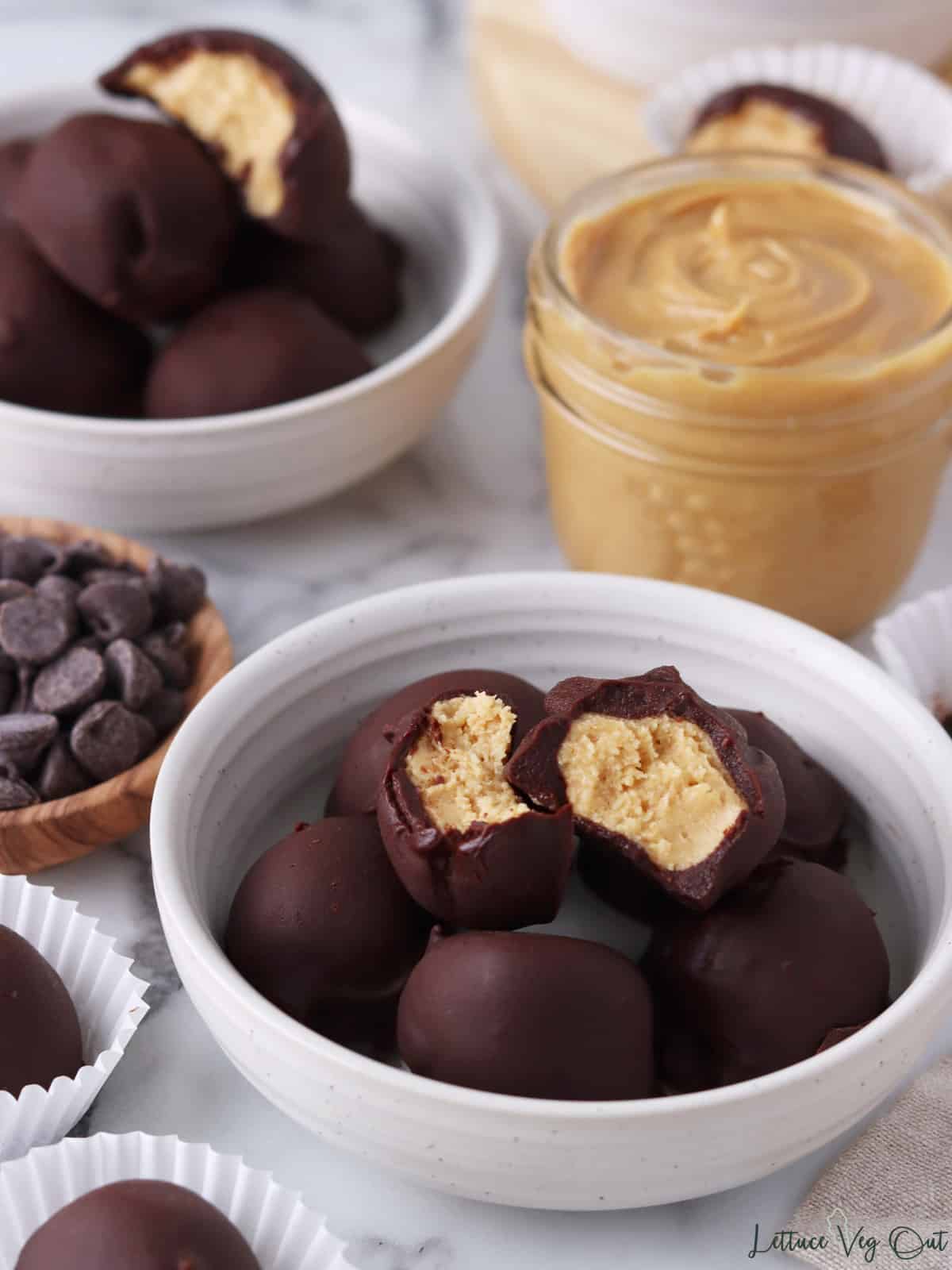 Two dishes of peanut butter balls with bites taken from some, along with a jar of peanut butter and small wood dish of chocolate chips.