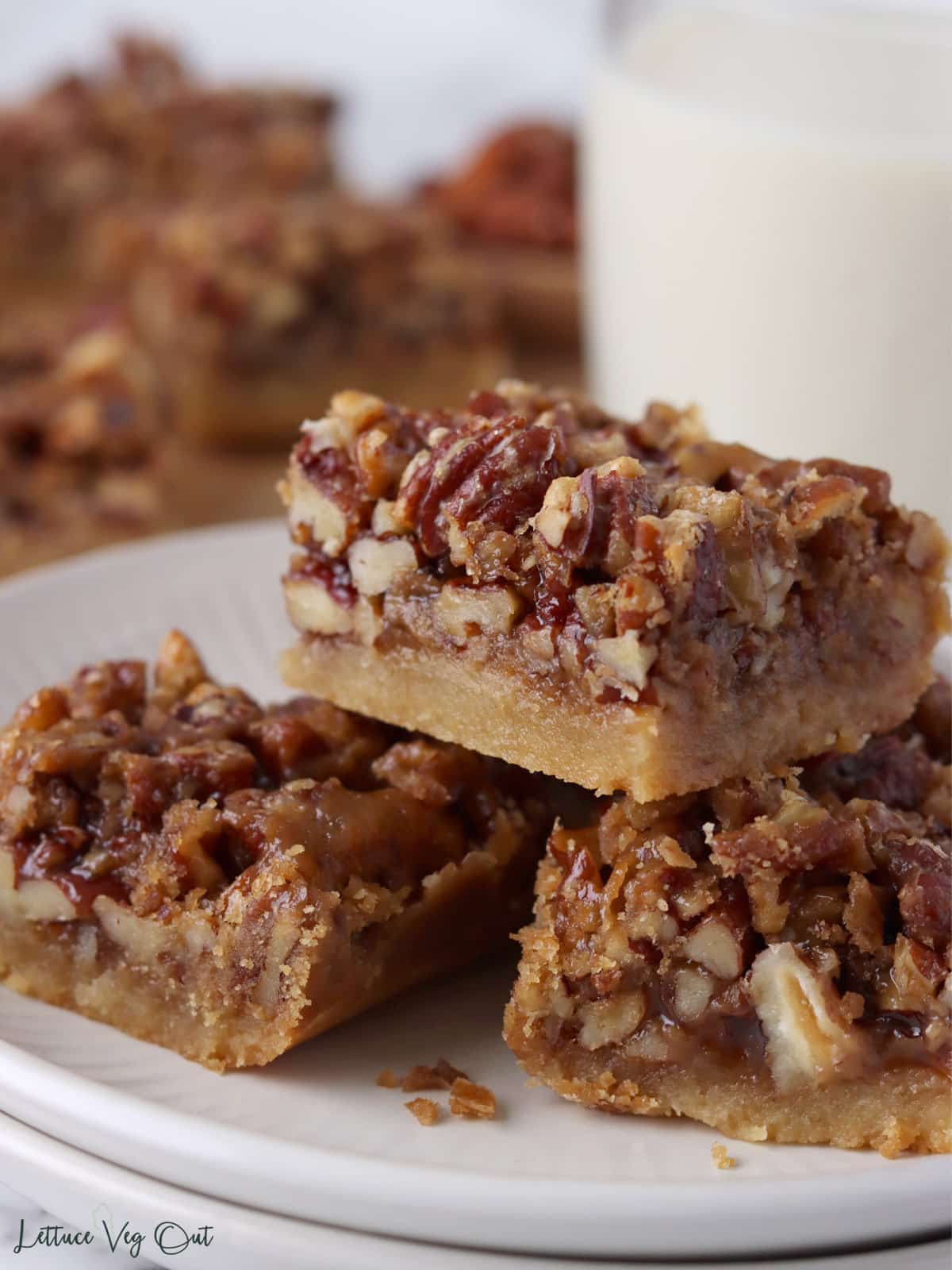 Stack of three pecan pie squares on a plate with a blurred glass of milk and board of more squares in back.