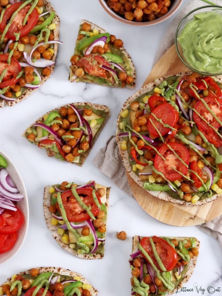 Arrangement of whole pizzas and pizza slices that are topped with vegetables, BBQ chickpeas and green sauce.