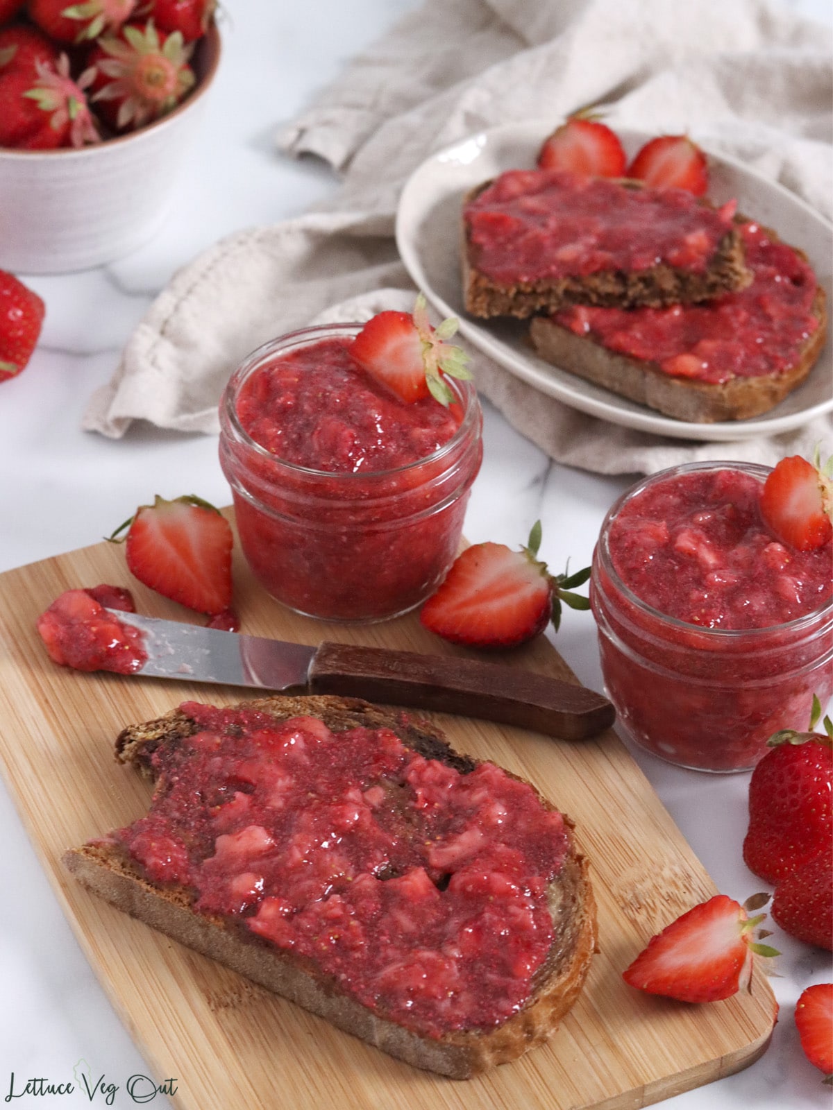 Toast covered with strawberry jam on wood board with small jars of jam and a small knife.