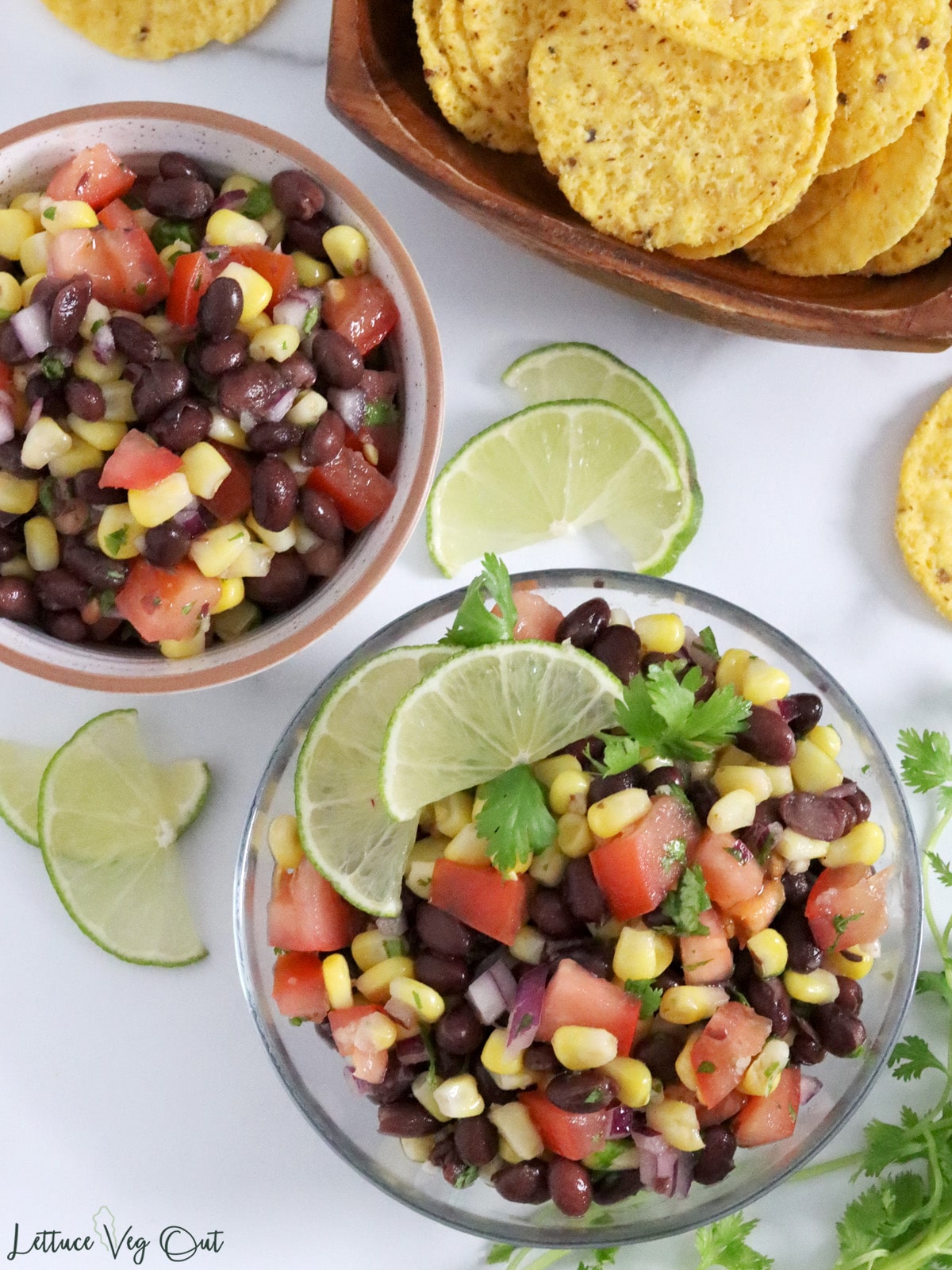 Two bowls of corn and bean salsa along with a bowl of tortilla chips and lime wedge garnishes.
