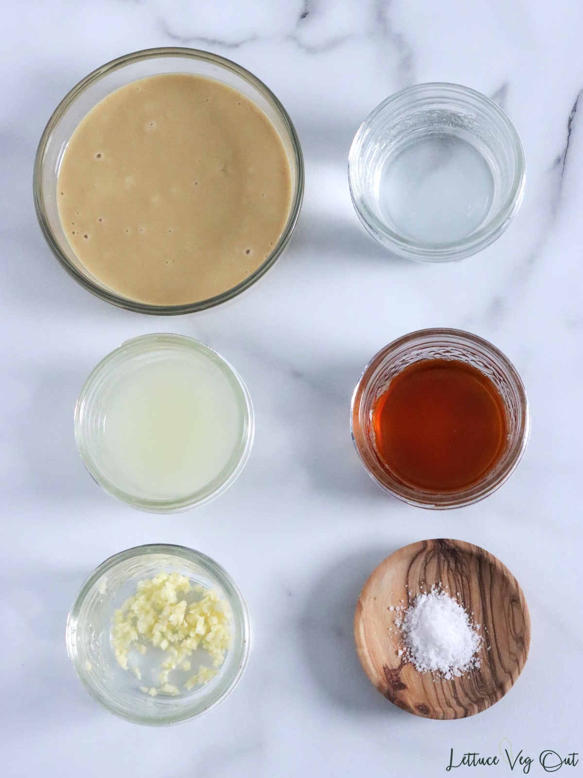 Ingredients for tahini sauce in glass dishes (tahini, water, lemon juice, maple syrup, minced garlic, salt).