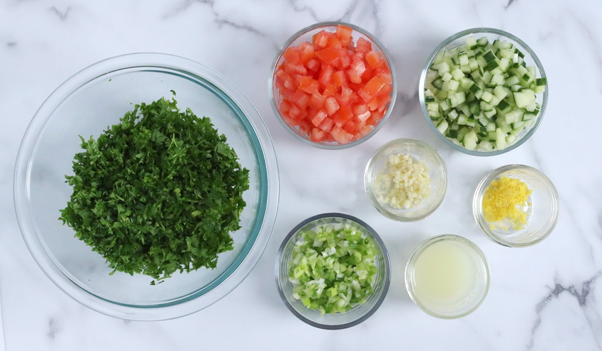 Small bowls of chopped tabbouleh ingredients (parsley, tomato, cucumber, garlic, green onion, lemon zest, lemon juice).