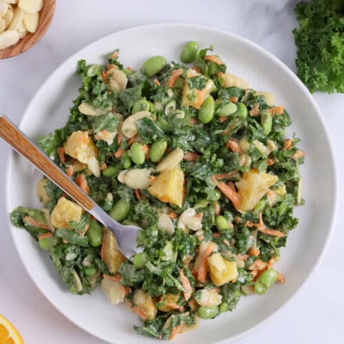 Close up of a plate of kale salad with tahini dressing, oranges, edamame, almonds and carrots.