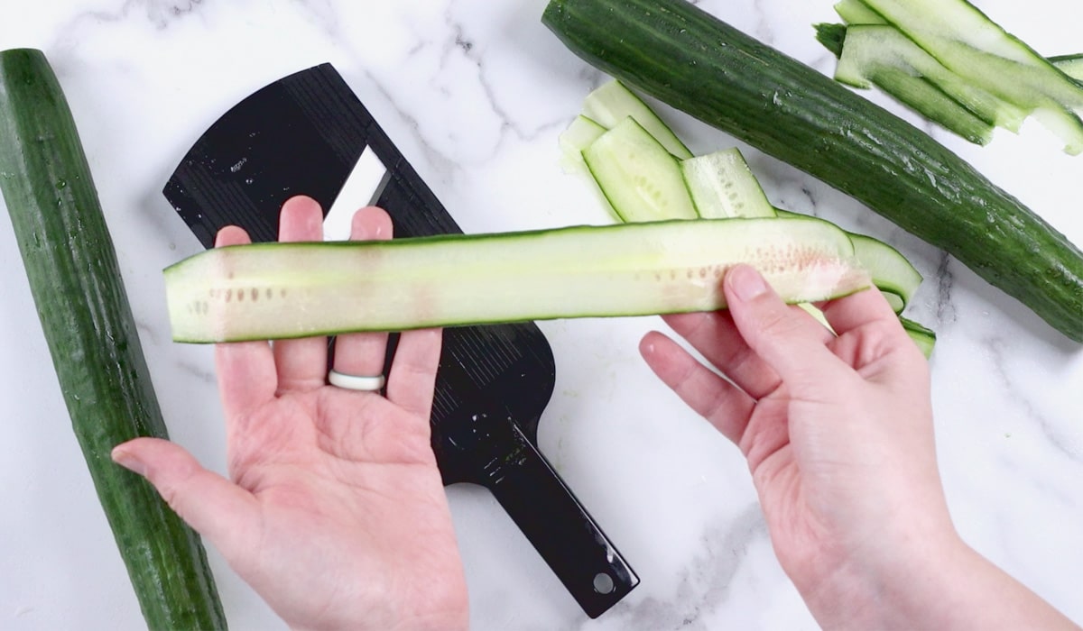 Hands holding a long, thin slice of cucumber.