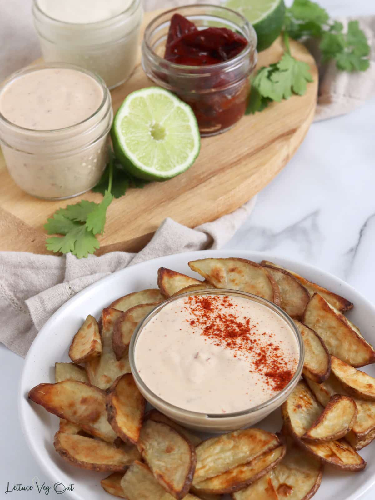 Plate of potato wedges with chipotle mayo sauce in front of wood board with chipotle peppers, more chipotle mayo, lime slices and cilantro.