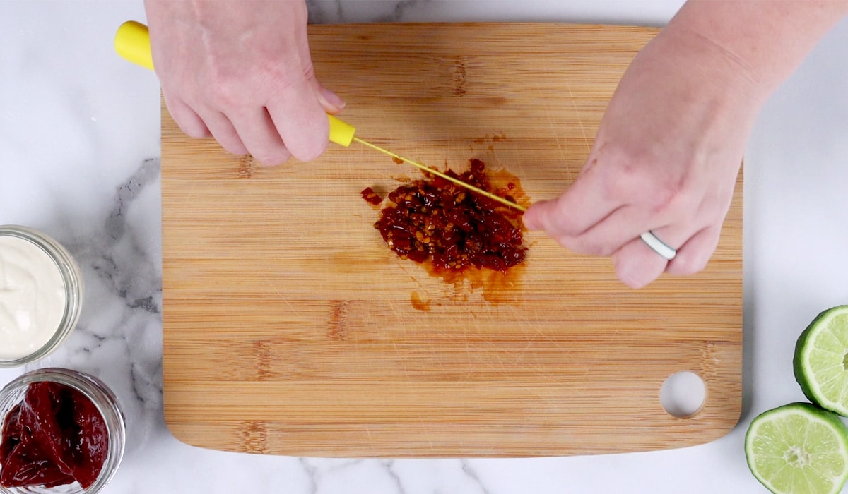Hands chopping chipotle peppers on a wood board.