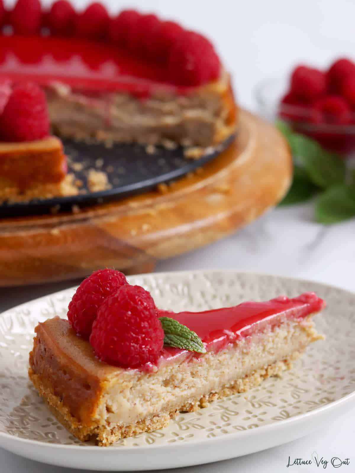 Close up of a slice of tofu cheesecake with raspberry topping and blurred cake in background.