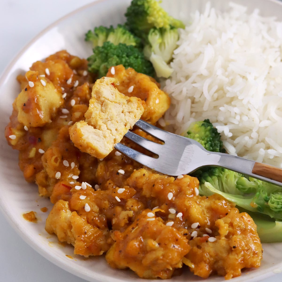 Close up of a bite of orange tofu on a fork over more orange tofu, broccoli and rice.