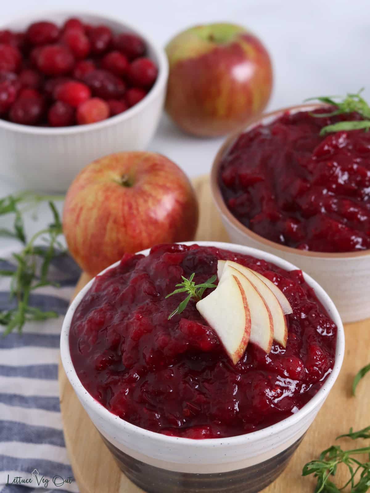 Two bowls of cranberry sauce on wood board with apple and herb garnish and bowl of whole cranberries in back.