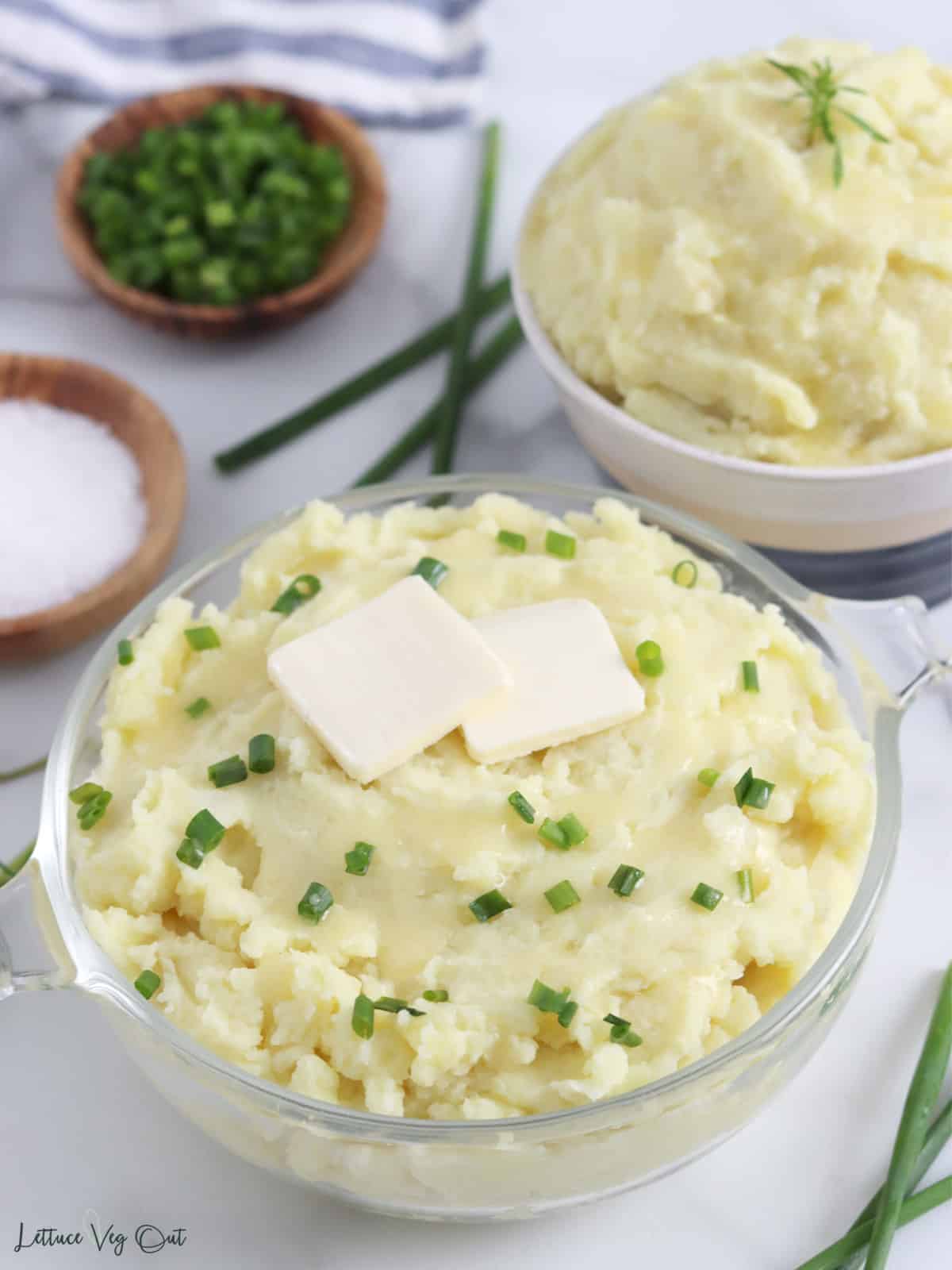 Glass bowl filled with creamy mashed potatoes topped with pats of vegan butter and chives with second bowl of mash in background.
