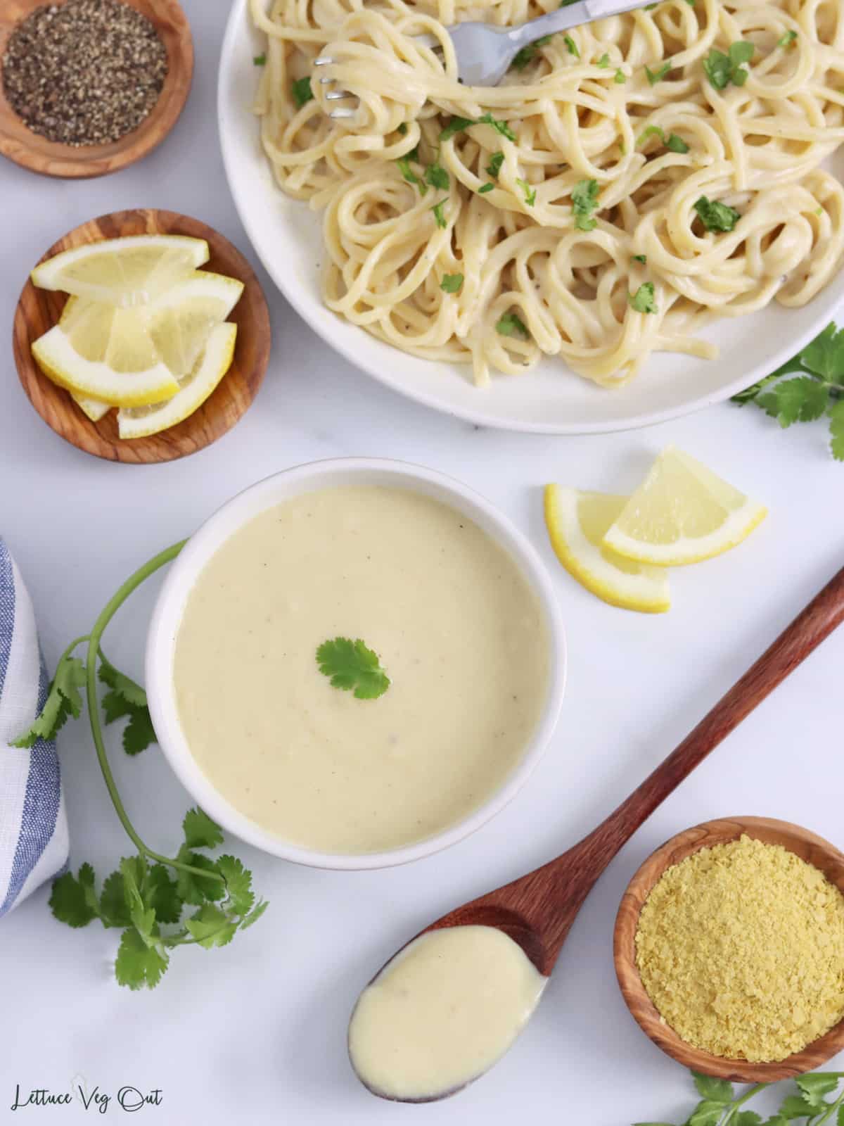 Bowl of white sauce with spoonful of sauce, lemon slices, and a plate of pasta tossed in bechamel surrounding the bowl.