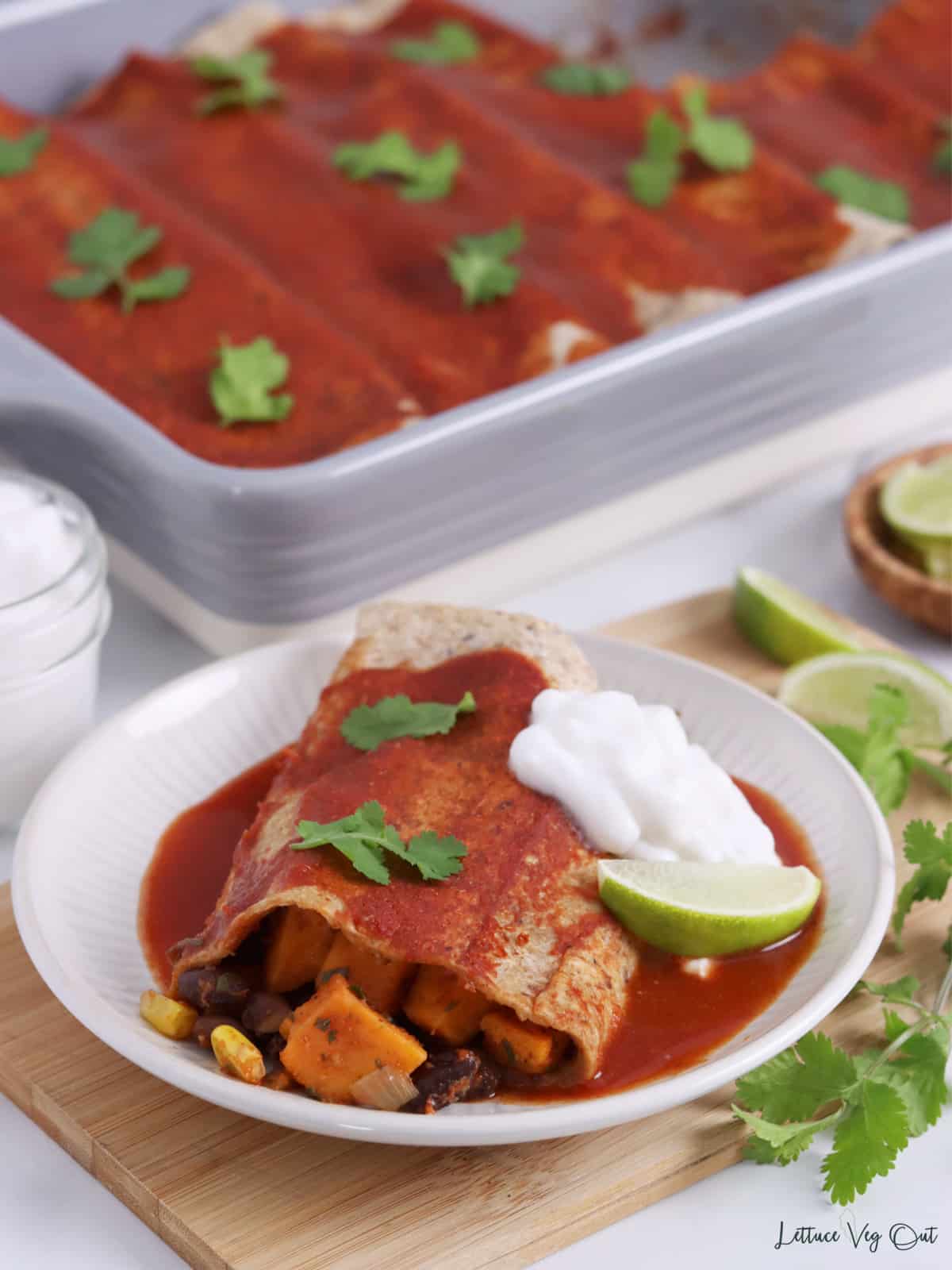 Close up of a cut open black bean enchilada with red sauce and sour cream with baking dish of enchiladas in background.