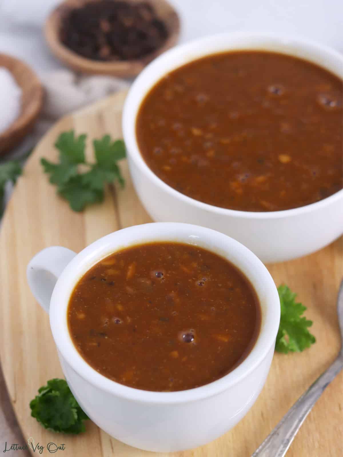 A small cup and bowl of brown gravy on a wood board.